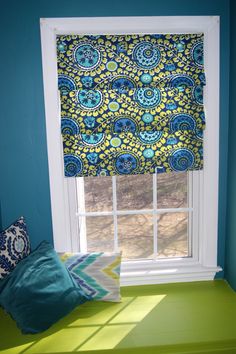 a window with blue and green curtains in the corner next to a pillow on a bench