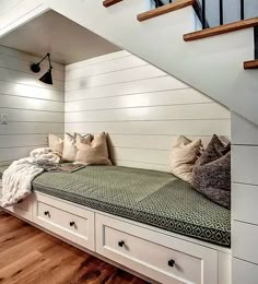 a bench with pillows and blankets on it under the stairs in a room that has wood flooring