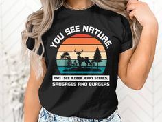 "Embrace your love for the great outdoors and your sense of humor with this Nature Deer Graphic Tee! The shirt displays a striking image of a majestic deer amidst a forest landscape, overlayed by the colors of a serene sunset. Above, the whimsical text, \"YOU SEE NATURE,\" is contrasted with the punchline at the bottom, \"AND I SEE A DEER JERKY STEAKS, SAUSAGES AND BURGERS\", making for a funny twist that is sure to be a conversation starter at any BBQ or camping trip. Made with high-quality material, this T-Shirt is perfect for anyone who appreciates wildlife and a good laugh. It's a comfortable, unisex fit that's ideal for casual everyday wear or as a special gift for the camping enthusiast or foodie in your life. Product Features: Fabrication: 100% Airlume combed and ring-spun cotton, 3 Funny Letter Print T-shirt For Outdoor, Outdoor Funny Print Crew Neck Tops, Outdoor Crew Neck Tops With Funny Print, Funny Print Crew Neck Top For Outdoor, Funny Text Short Sleeve T-shirt For Outdoor, Casual Outdoor T-shirt With Funny Text, Deer Jerky, Shirt Displays, Majestic Deer