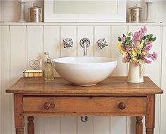 a bathroom sink sitting on top of a wooden table next to a mirror and vase with flowers