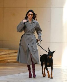 a woman in a trench coat walking her dog on a leash through the city streets