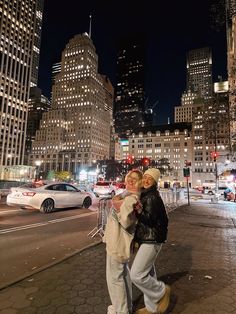 two people standing in the middle of a city street at night with tall buildings behind them