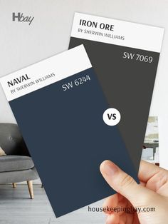 The image compares two dark paint swatches. On the left is "Naval" by Sherwin Williams with the code SW 6244, a deep navy blue with strong, rich undertones. On the right is "Iron Ore" by Sherwin Williams with the code SW 7069, a dark charcoal gray with warm undertones. A hand is holding both swatches, and a "VS" symbol in the middle indicates a comparison. The background is blurred, featuring a neutral-toned room. These colors are perfect for making bold design statements in interior  spaces. Naval Sw 6244, Exterior Refresh, Deep Gray