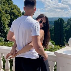 a man standing next to a woman on top of a white balcony with trees in the background