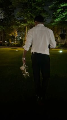 a man in white shirt and black pants walking on grass at night with trees behind him