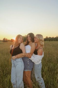 three women standing together in a field with their arms around each other as the sun sets