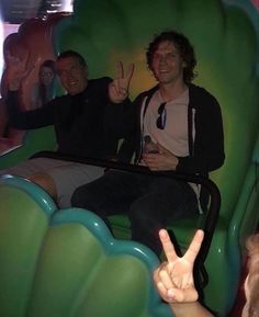 two men sitting in a green chair giving the peace sign with one hand and another pointing up