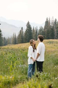 a man and woman standing in the grass with mountains in the background at their side