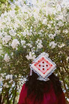 a woman in a red dress with a white square on her head and flowers behind her