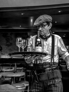 a man holding a tray with two wine glasses on it while wearing an apron and bow tie