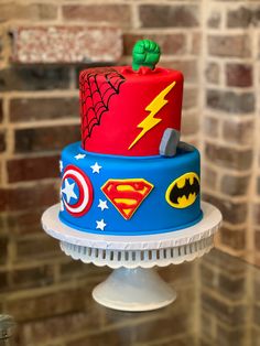 a birthday cake decorated with superman and captain america symbols on a pedestal in front of a brick wall