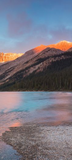 the mountains are covered in snow as the sun is setting on the water and rocks