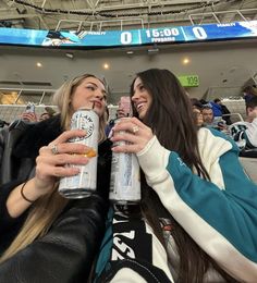 two women holding up cans of beer at an ice hockey game in front of the stands