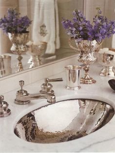 a bathroom sink sitting under a mirror with purple flowers in the vase next to it