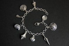 A collection of silver plated seashell themed charms have been dispersed around a shimmering silver plated bracelet chain in this handmade charm bracelet. This sea shell charm bracelet is then completed with a lobster clasp and a 1/2 inch of chain at the end for adjustable sizing. Charms in this bracelet include a starfish on a scallop shell charm, sand dollar charm, two scallop shell charms, conch shell charm, mussel shell charm, sea snail shell charm, long spiral shell charm, and spindle shell Silver Shell Bracelets Gift, Silver Shell-shaped Pendant With Lobster Clasp, Silver Metal Charm Bracelet For Beach, Silver Charm Bracelet With Lobster Clasp For Beach, Silver Charm Bracelet For The Beach, Handmade Silver Charm Bracelet For Beach, Sea Snail Shell, Handmade Charm Bracelets, Seashell Bracelet