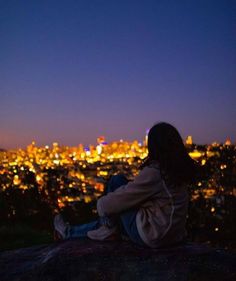 a woman sitting on top of a rock looking at the city lights in the distance