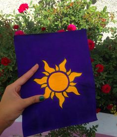 a hand holding up a purple and yellow square with the sun painted on it in front of red flowers