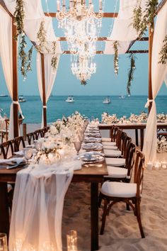 a dining table set up on the beach for an outdoor dinner party with chandelier and candles