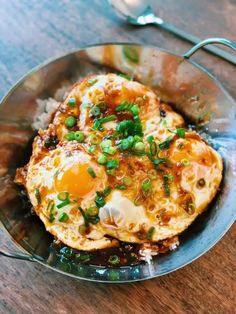 two fried eggs are on top of rice in a metal bowl with a spoon and fork next to it