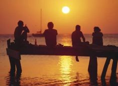 four people sitting on a dock watching the sun go down over the ocean at sunset