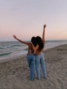 two women are standing on the beach with their arms in the air and facing the ocean