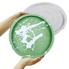 two hands holding green plates with white paint splattered on them, and one person's hand reaching for the plate