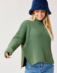 a woman in a green sweater and blue hat smiles while holding her hands out to the side