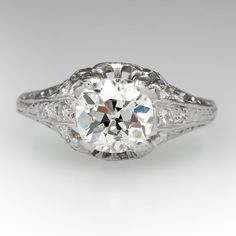 This ring features a pierced design and accented with engraved wheat motifs. The ring is centered with one (1) old mine cut diamond set with four- prongs. The shoulders of the ring are each accented with four (4), bead set, round single cut diamonds. The ring measures 8.7mm at the top, rises 5.9mm above the finger, tapering to 1.4mm wide and 0.9mm thick at the base of the shank. Antique Cushion Cut Anniversary Rings, Heirloom Diamond Ring With Cushion Cut, Classic Round Engraved Ring With Center Stone, Classic Engraved Ring With Center Stone, Classic Engraved Platinum Ring, Antique Round Cut Diamond Ring, Classic Platinum Engraved Ring With Round Cut, Classic White Filigree Ring With Brilliant Cut, Heirloom White Gold Diamond Ring With Classic Cut