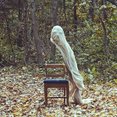 a ghost sitting on top of a wooden chair in the middle of a leaf covered forest