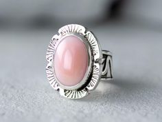 Bold but dainty Pink Conch Silver ring on a unique silver band that has been handmade using scrap 925 Sterling Silver. The band has been fused, oxidized then lightly polished. At its widest the band is 3/8" then tapers to 1/8". The oval cabochon is 14mm x 10mm and is in a bezel setting accented with stamped, cut out square wire. Add a touch of Coastal Cowgirl attitude to your outfit with our simple, but elegant Pink Conch Silver Statement Ring. Whether you're shopping for yourself or are searchi Pink Oval Opal Ring In Sterling Silver, Pink Opal Sterling Silver Ring Gift, Silver Pink Opal Ring For Gift, Pink Opal Ring In Sterling Silver For Gift, Silver Pink Opal Ring Gift, Pink Opal Rings As A Gift, Adjustable Pink Opal Ring As A Gift, Handmade Sterling Silver Pink Rings, Handmade Pink Sterling Silver Rings