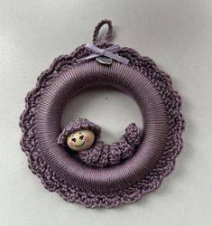 a purple knitted wreath with a small doll hanging from the front, on a white surface