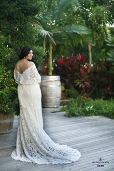 a woman in a white dress is standing on a wooden deck near some bushes and trees