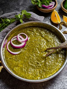 a metal bowl filled with green sauce and onions on top of a gray surface next to sliced oranges