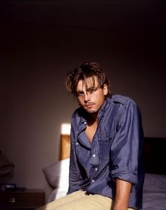 a man sitting on top of a bed next to a white wall and wearing a blue shirt