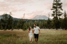 a man and woman holding hands walking through a field with mountains in the back ground