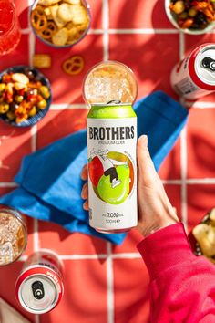 a person holding up a can of brothers apple cider next to bowls of food