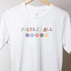 Introducing our new pickleball shirt, perfect for both on and off the court! Made from a lightweight fabric, this shirt will keep you comfortable no matter how long your pickleball game lasts. The relaxed fit ensures that you have full range of motion, allowing you to move freely and play your best. This shirt features a classic crew neck design and short sleeves, making it versatile enough to wear on any occasion. The shirt also features a fun and colorful design with a pickleball theme, showing off your love for the sport. The high-quality print will stay vibrant even after many washes, ensuring that you can enjoy wearing this shirt for years to come. This casual pickleball shirt is available in a range of sizes to fit every body type. It's perfect for both men and women and can be dress Fun White Sports T-shirt, White Moisture-wicking Tennis T-shirt, White Moisture-wicking T-shirt For Tennis, White T-shirt With Team Name For Pickleball, Sporty White T-shirt For Pickleball, Pickleball Shirts, Pickleball Shirt, Pickleball Gift, Custom Shirt