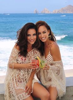 two beautiful women sitting next to each other near the ocean holding drinks and posing for a photo