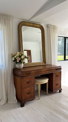 a wooden desk with a mirror and stool in front of a white curtained window