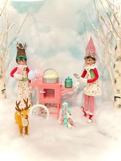 two dolls are standing in the snow near a table with food and a toy deer