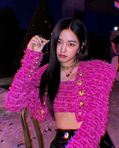 a woman with long black hair sitting on a chair wearing a pink top and gold jewelry