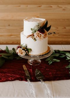 a white wedding cake with flowers and greenery on a red table cloth next to silverware