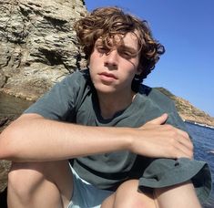 a young man sitting on top of a rock next to the ocean with his arms crossed