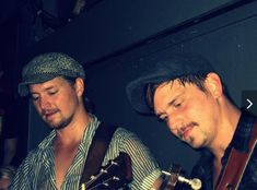 two young men are playing guitars in a dark room, one is wearing a hat and the other wears a striped shirt