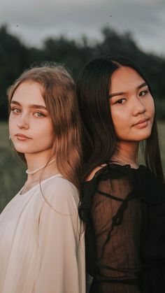 two women standing next to each other in front of a field with grass and trees