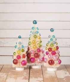 three small christmas trees with colorful ornaments on them sitting on a wooden table in front of a white wall