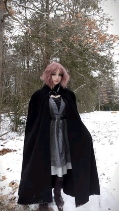 a woman with pink hair wearing a black cape and dress in the snow near some trees