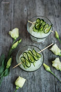 two glasses filled with cucumber on top of a wooden table next to flowers