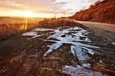 the sun is setting over an empty road with puddles on it and grass in the foreground