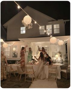 a group of women standing around a table in front of a house with lights hanging from the ceiling